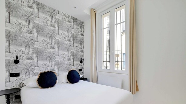A bedroom with a large bed, a patterned wallpaper, a window with curtains, and a nightstand.