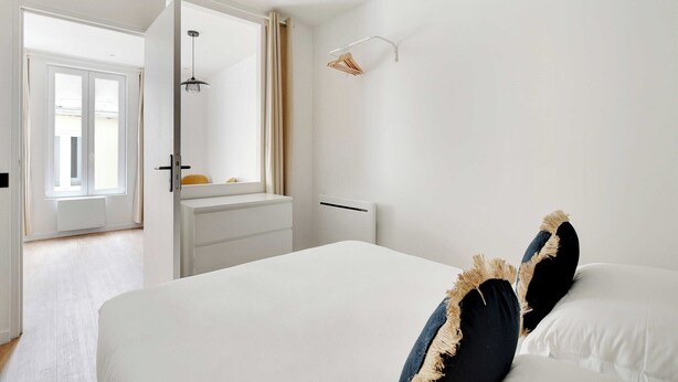 Une chambre à coucher comprenant un lit, une table de chevet, un luminaire suspendu, un radiateur et une fenêtre avec des rideaux.