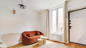 Un salon moderne comprenant un canapé, un fauteuil blanc, deux petites tables rondes, un luminaire suspendu et une grande fenêtre avec des rideaux.