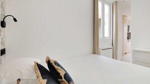 Makuuhuoneessa on sänky, seinään kiinnitetty valaisin, ikkuna, jossa on verhot, ja näkymä toiseen huoneeseen.