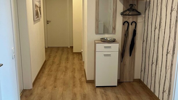 Un couloir doté d’un plancher en bois, d’une armoire blanche avec des tiroirs, d’un miroir et d’un porte-manteau avec deux parapluies.