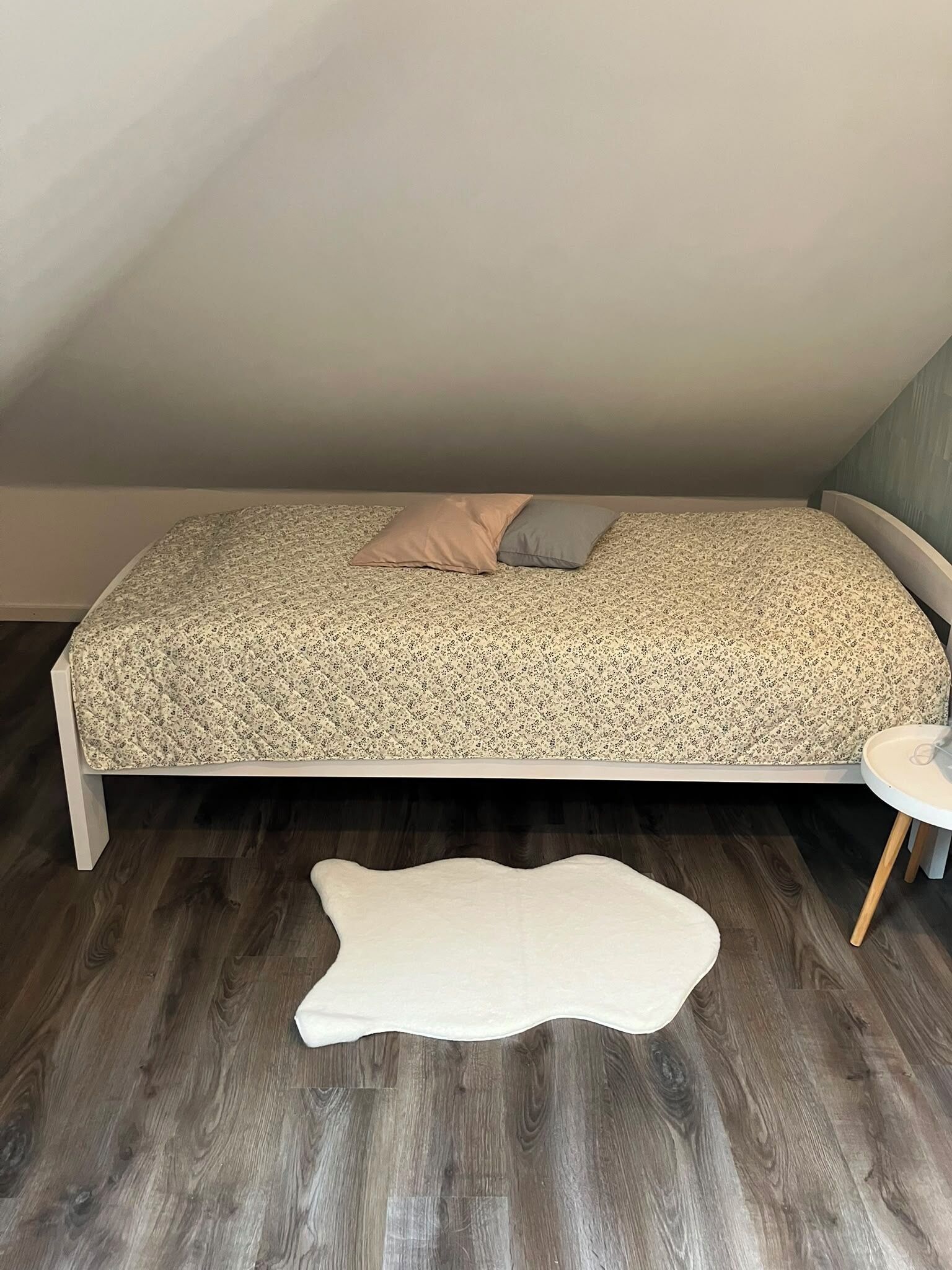 Een slaapkamer met een bed, een klein rond tafeltje en een witte kussen op de vloer.