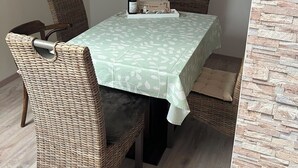 Un coin repas avec une table recouverte d’une nappe à motifs de feuilles, des chaises en osier, une bouteille de vin et une petite boîte décorative.
