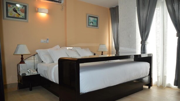 A bedroom with a bed, two bedside tables, a lamp, and a wall-mounted air conditioner. - Saint David Hôtel Résidence (Douala)