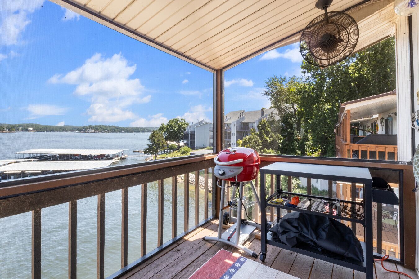 A balcony with a grill, a fan, and a view of a lake and buildings.