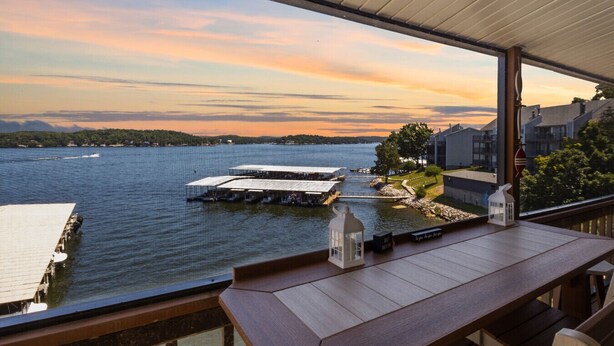 A lakeside view from a covered deck with a wooden table and chairs, overlooking a dock with boats and a small island with buildings.