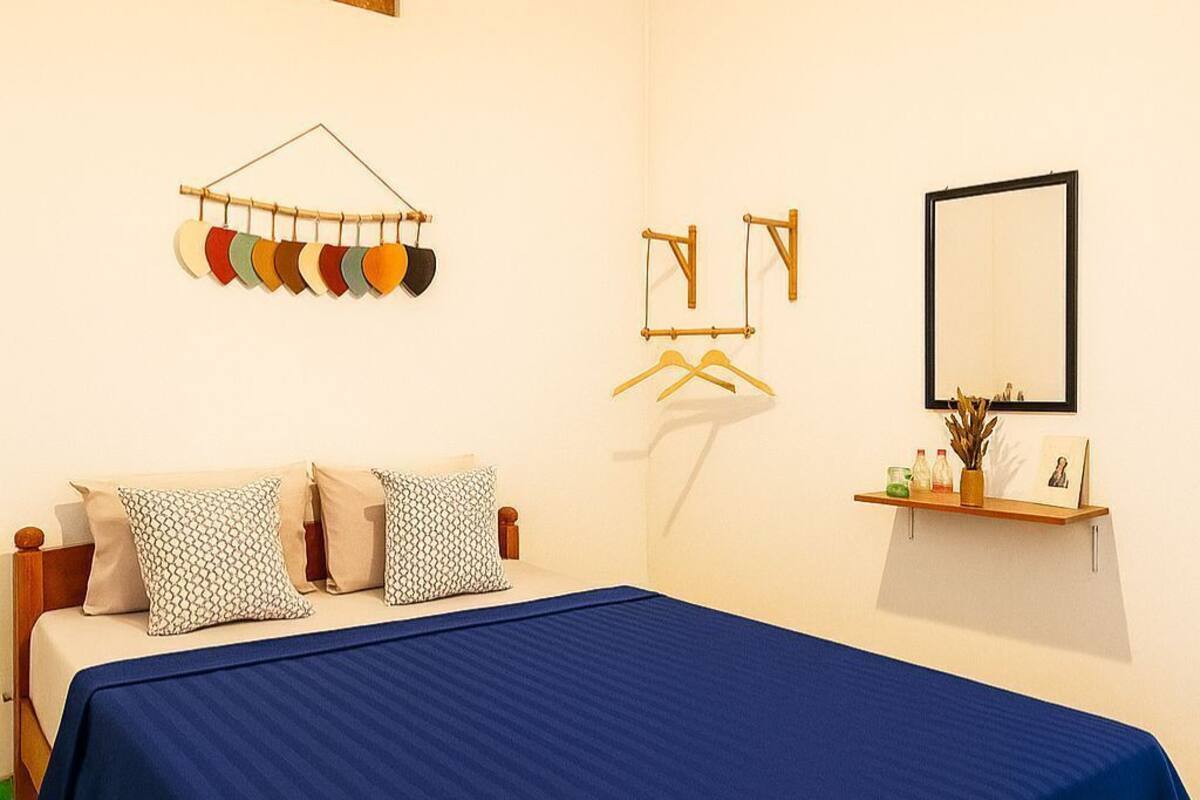 A bedroom with a bed, a hanging light fixture, a wall-mounted mirror, and a shelf with items.