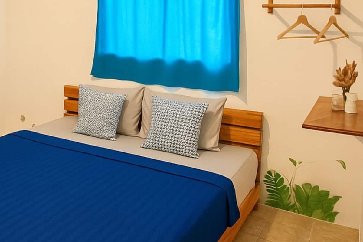 A bedroom with a bed, blue curtains, wooden headboard, two pillows, a bedside table, a small plant, and a wall-mounted shelf with a vase.