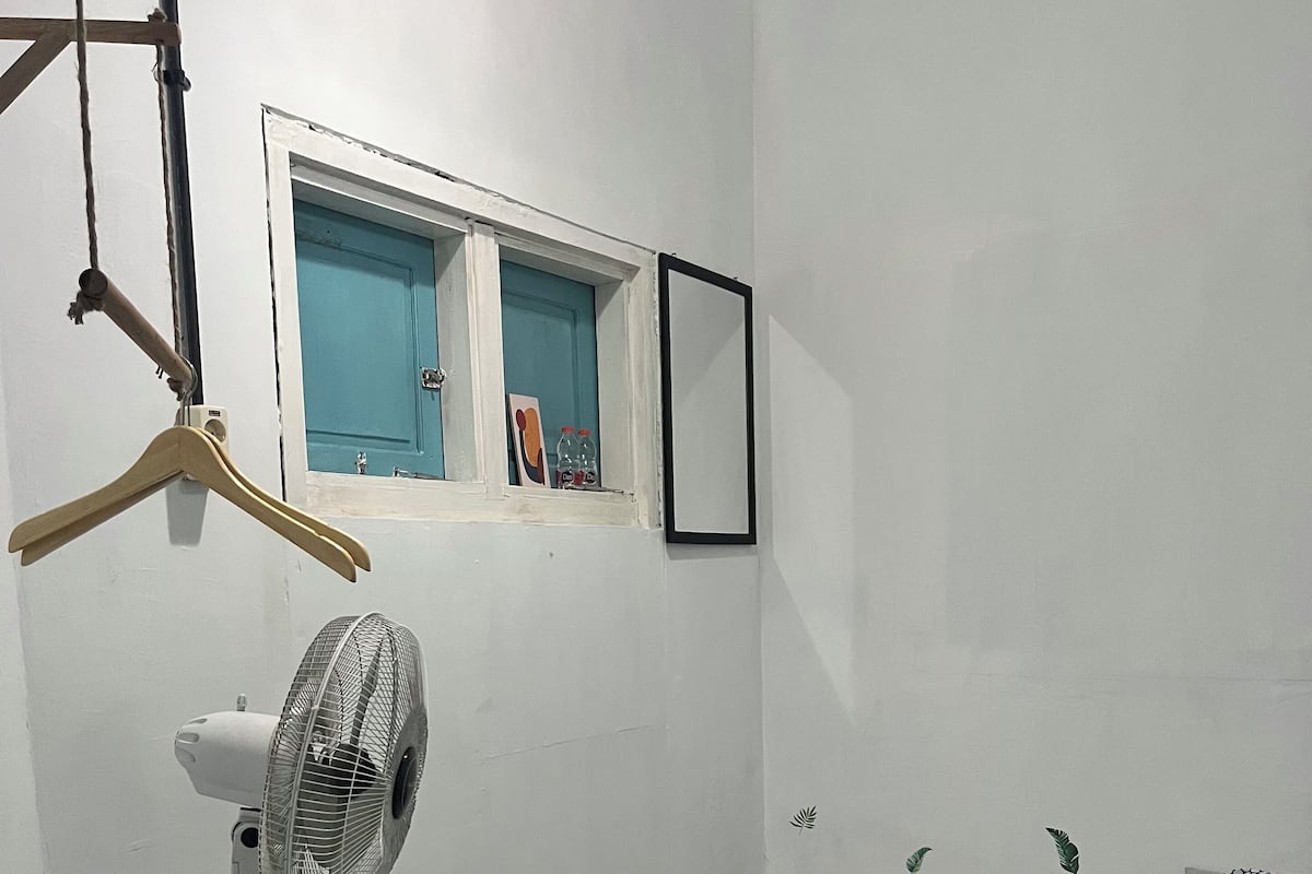 A room with a bed, a standing fan, a window with a view, and a wall-mounted mirror.