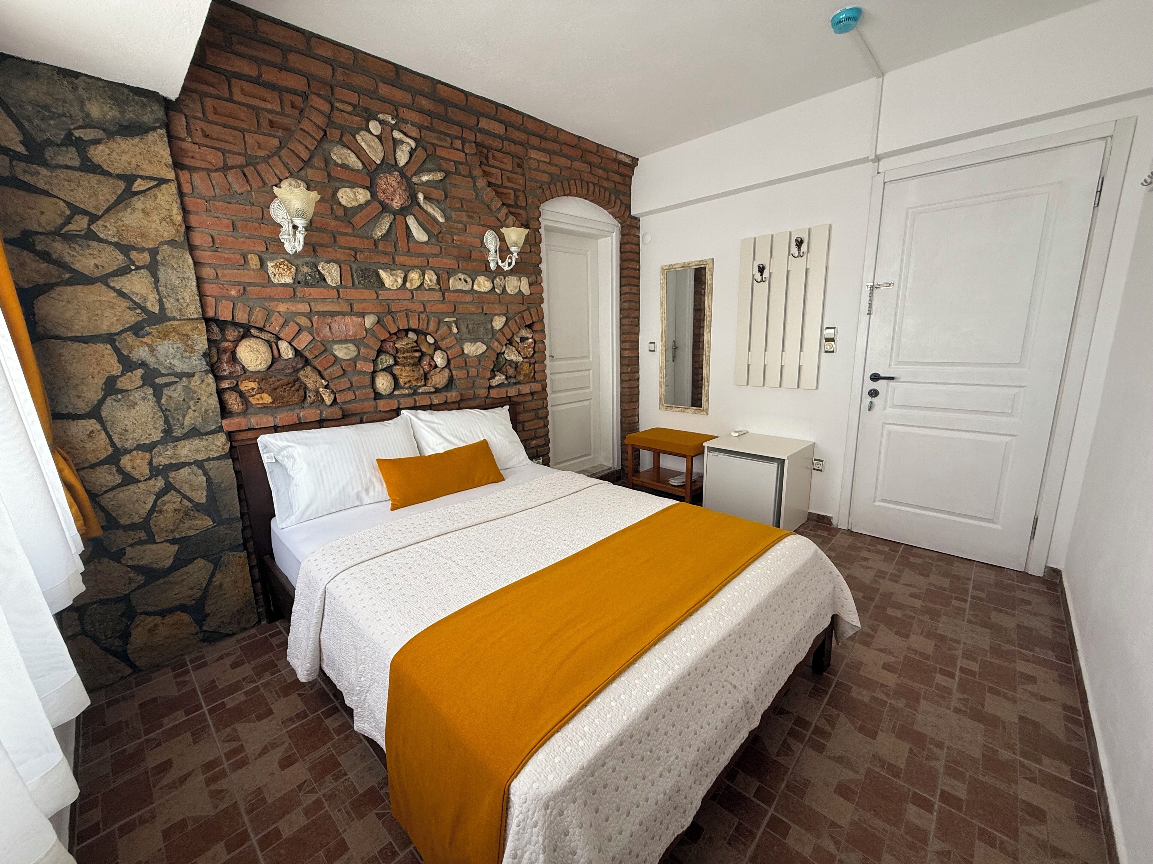 Un dormitorio con pared de ladrillo, cama con ropa de cama blanca y una manta mostaza, una mesita, un espejo y una puerta blanca.
