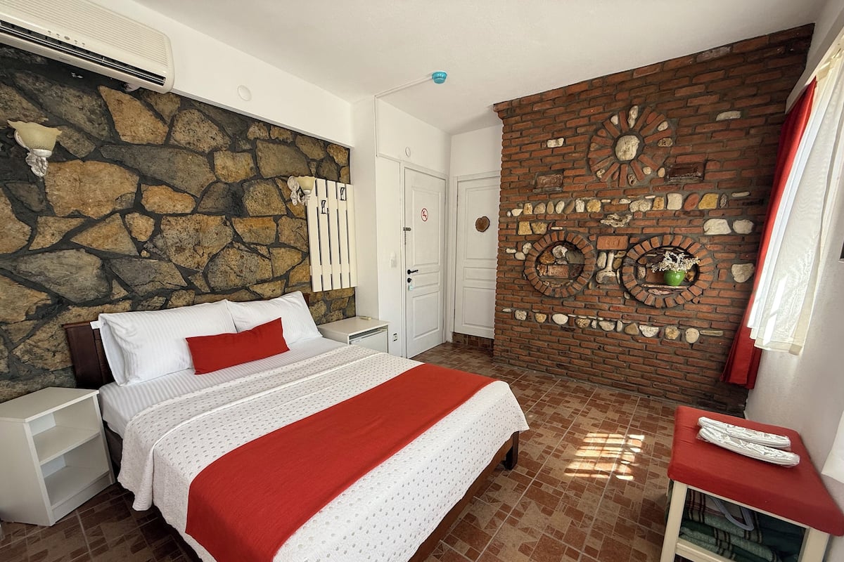 Um quarto com parede de pedra, lareira de tijolos, cama com roupa de cama branca e manta vermelha, e uma mesinha com almofada vermelha.