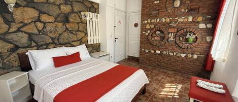 A bedroom with a stone wall, a brick fireplace, a bed with white bedding and a red throw, and a small table with a red cushion.