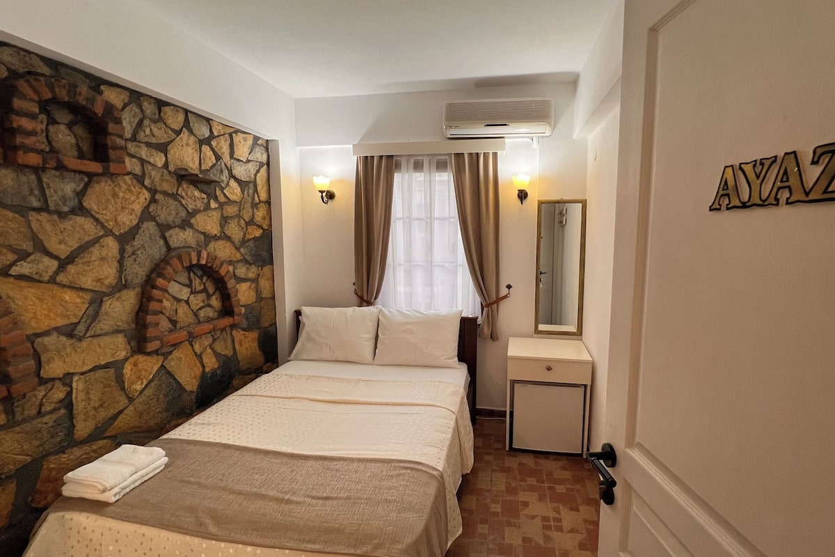 Quarto de hotel com parede de pedra, cama com roupa de cama branca, mesa de cabeceira, espelho e um letreiro com a inscrição "AYAZ".