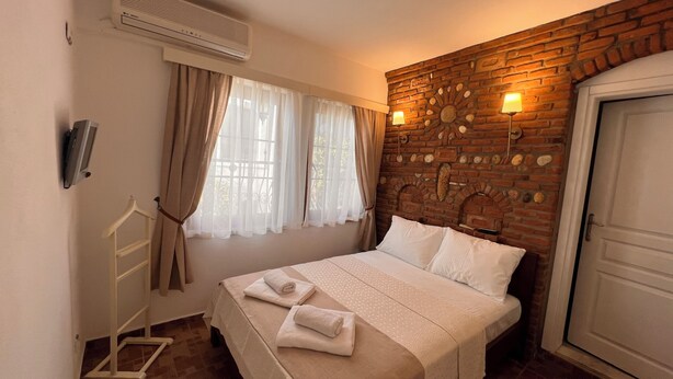 A hotel room with a bed, a window with curtains, a brick wall with a decorative element, a wall-mounted lamp, and a door. - Adahan Otel Bozcaada (Bozcaada)