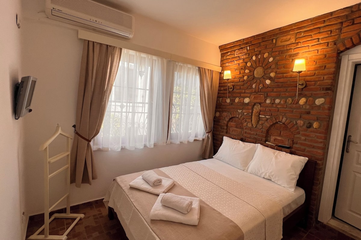 Quarto de hotel com cama, janela com cortinas, parede de tijolos com detalhe decorativo, luminária de parede e porta.
