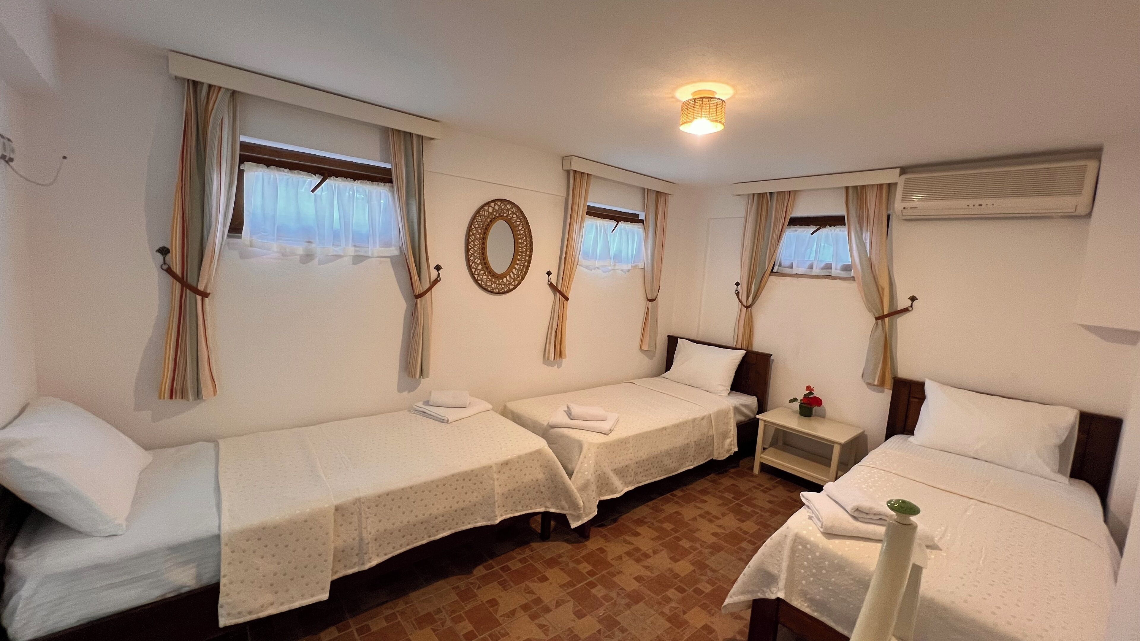 A hotel room with three single beds, a small bedside table with a lamp, and a window with curtains.