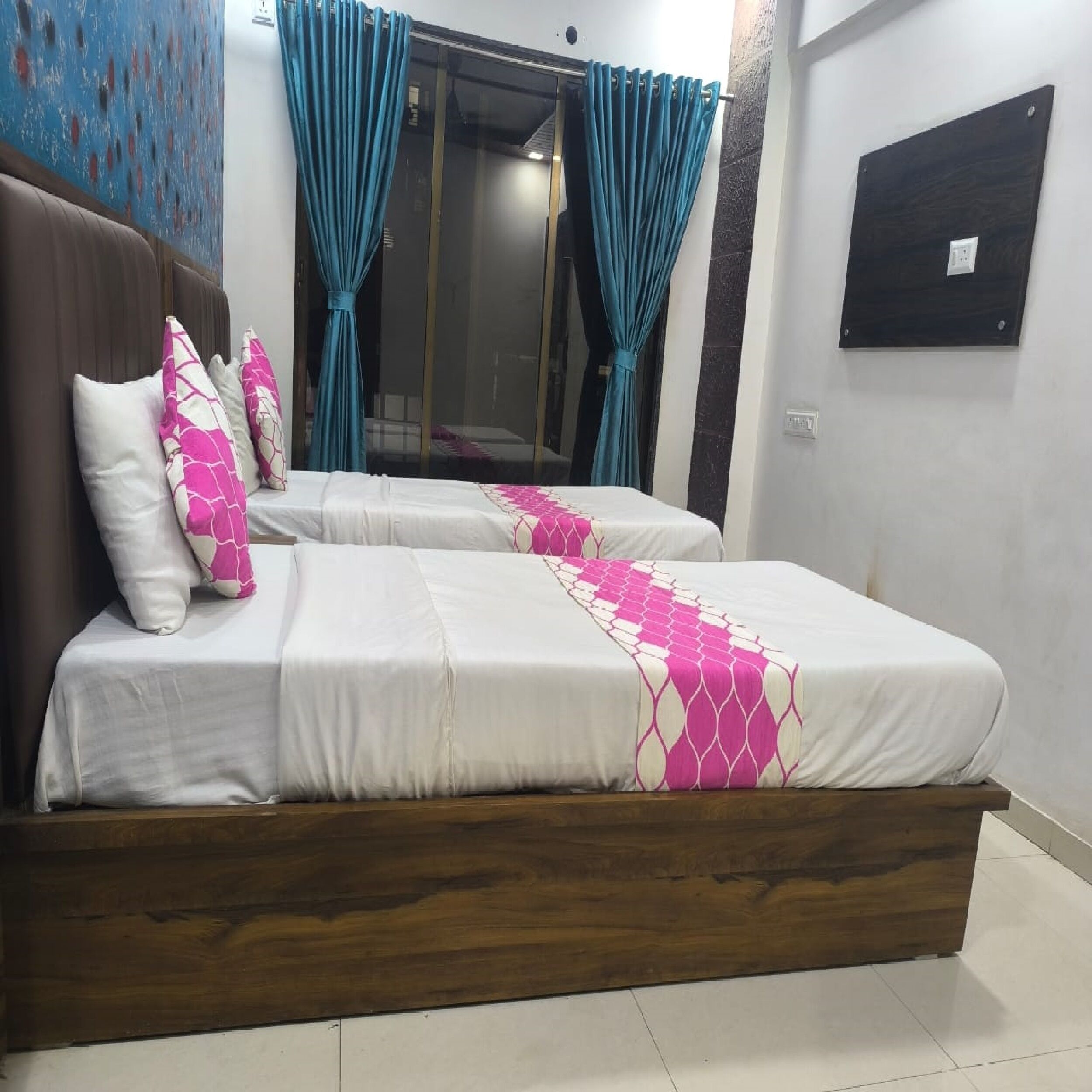 A bedroom with a wooden bed frame, two beds with white and pink bedding, teal curtains, and a wall-mounted light switch.