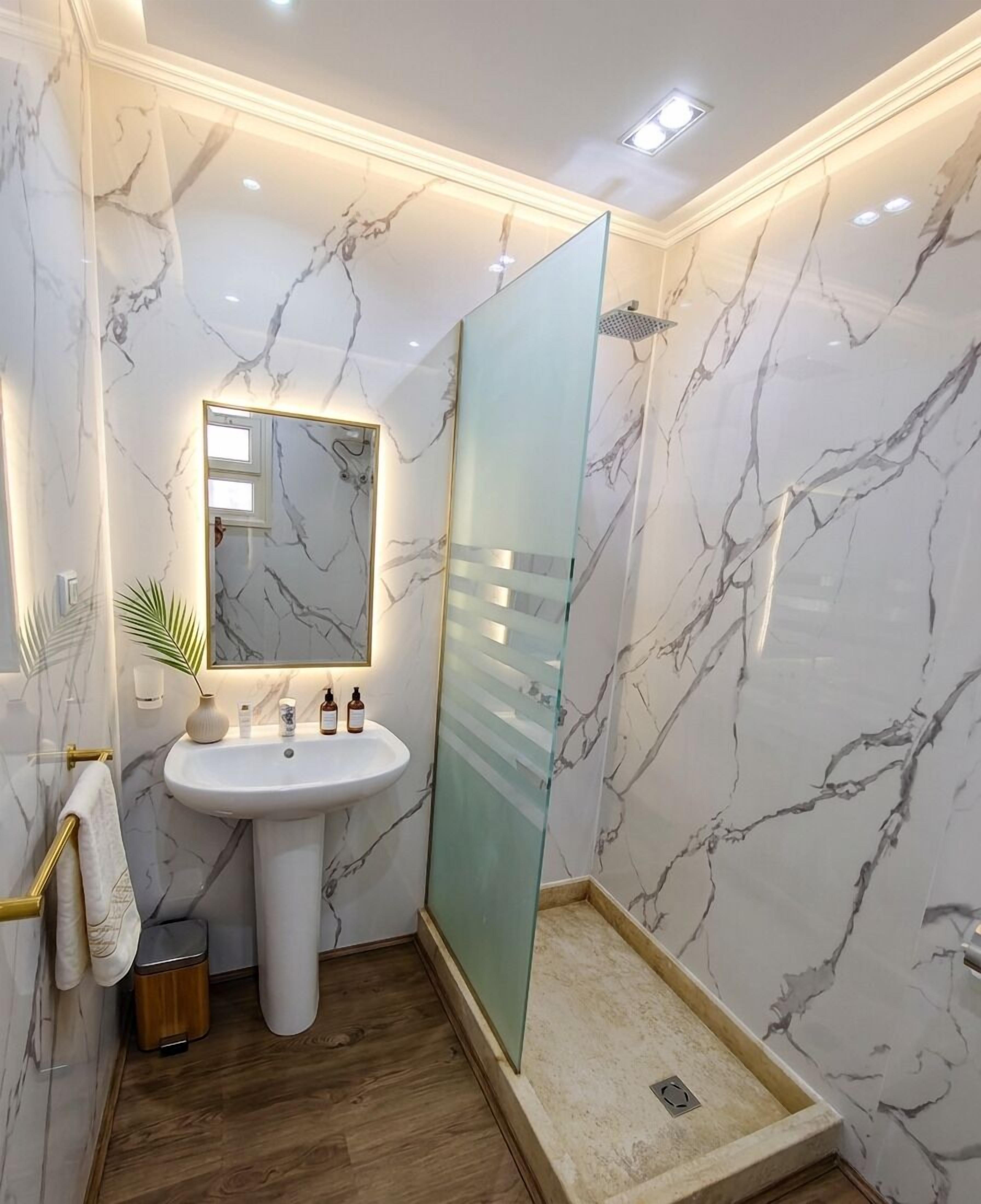 A bathroom with a marble wall, a pedestal sink, a glass shower enclosure, and a wooden floor.