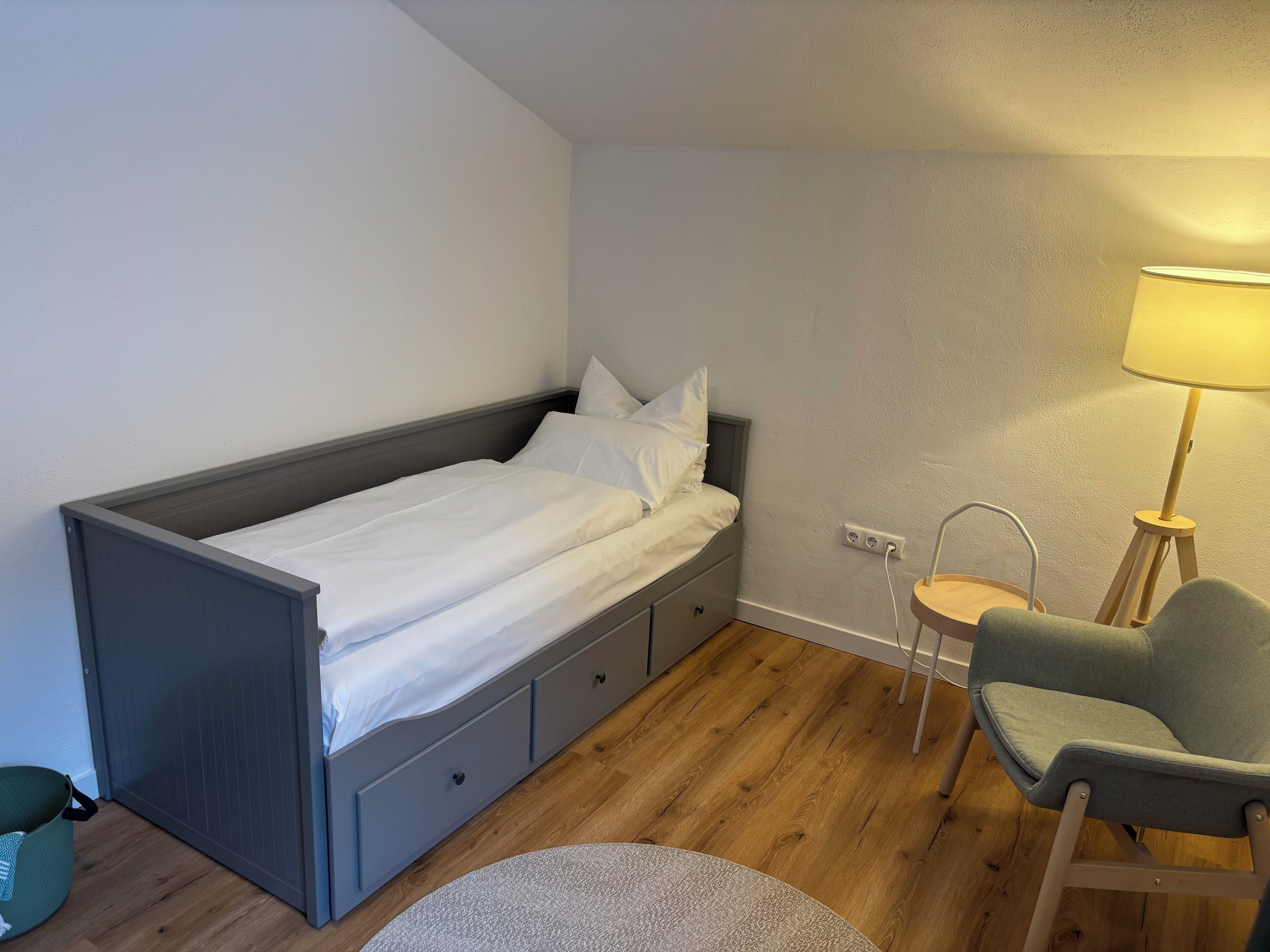 A small, single-bed room with a grey bed, a green chair, a small round table, and a floor lamp.