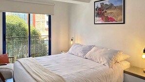 Un dormitorio con una cama grande, una mesita de noche, una lámpara de pared y un ventanal con vistas a edificios y árboles.