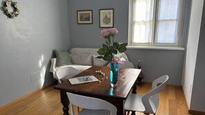 A dining area with a wooden table, white chairs, a vase with flowers, a framed picture on the wall, and a window with curtains.