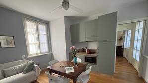 A modern living room with a wooden dining table, white chairs, a grey sofa, and a kitchen in the background.