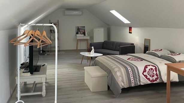 Une chambre moderne et bien éclairée, située sous les combles, avec un plafond en pente, un lit recouvert d’une literie à motifs, un canapé gris, une petite table et une armoire blanche avec des cintres en bois.