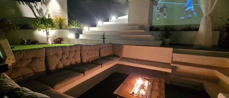 A cozy outdoor seating area with a fire feature, a TV screen displaying a soccer game, and a staircase leading to an upper level.