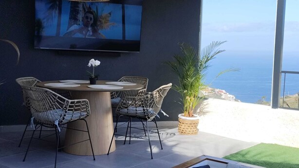 A modern outdoor dining area with a round table and wicker chairs, a potted plant, and a large TV screen on the wall. - Luxury Nest with Panoramic Wiews 10 min from Loro Park (Los Realejos)