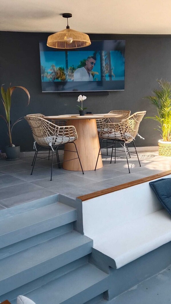 Un espace extérieur moderne, aménagé avec une table ronde en bois, deux fauteuils en osier et un téléviseur fixé au mur.