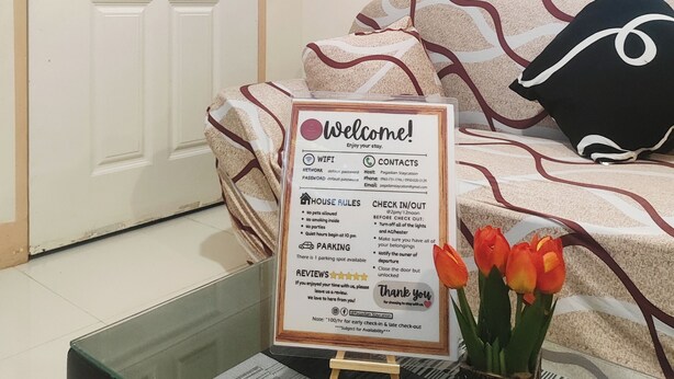 Habitación de hotel con una puerta blanca, un cartel de bienvenida con normas de la casa, una cama con ropa de cama estampada y un jarrón con tulipanes.