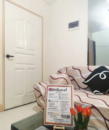 A hotel room with a white door, a welcome sign with house rules, a bed with patterned bedding, and a vase with tulips.