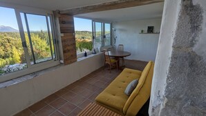 Un balcón con un sofá amarillo, una mesa de comedor y sillas. Amplias ventanas ofrecen vistas a un paisaje montañoso.