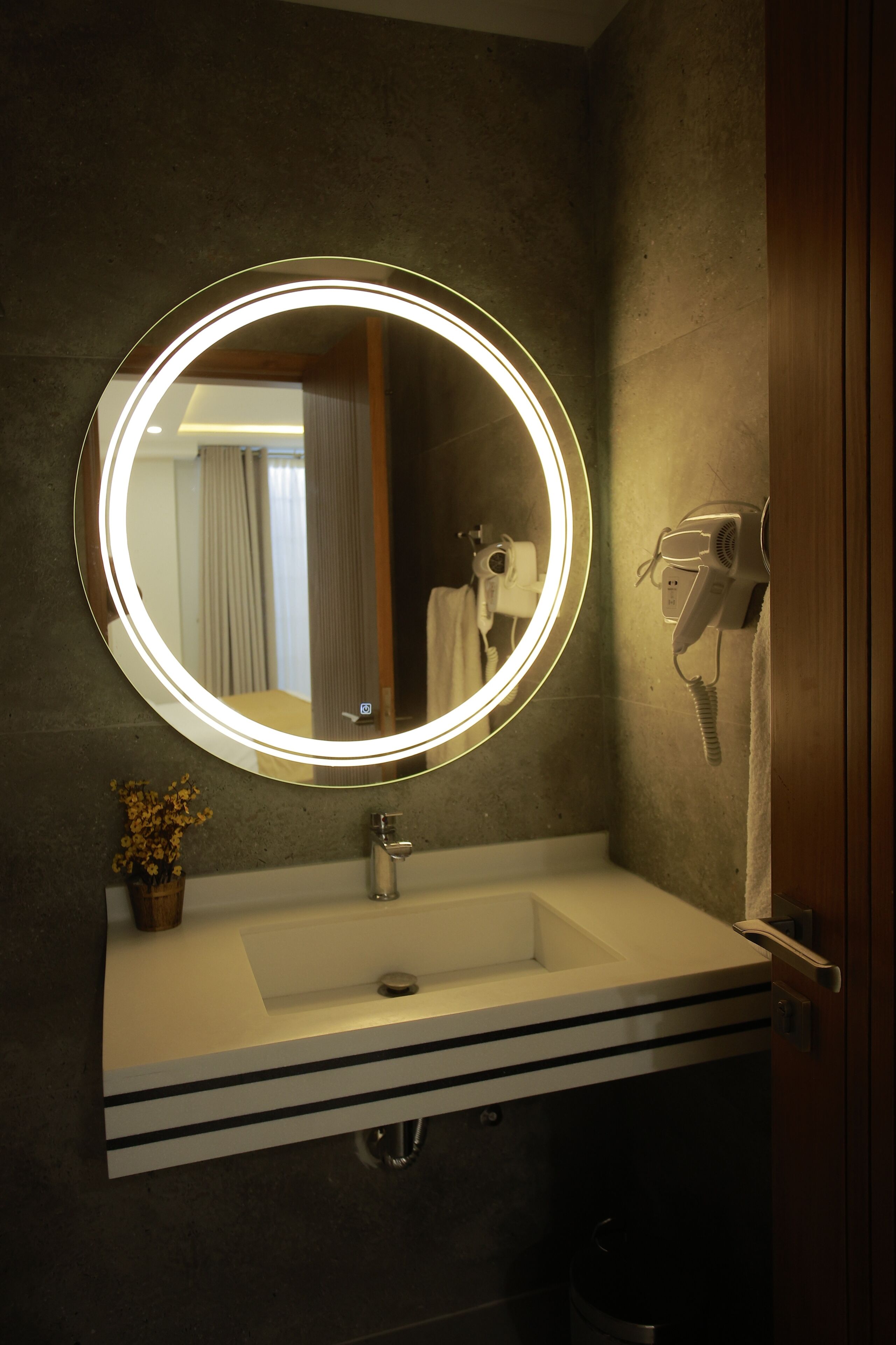 A bathroom with a round mirror, a sink, a hairdryer, and a trash bin.