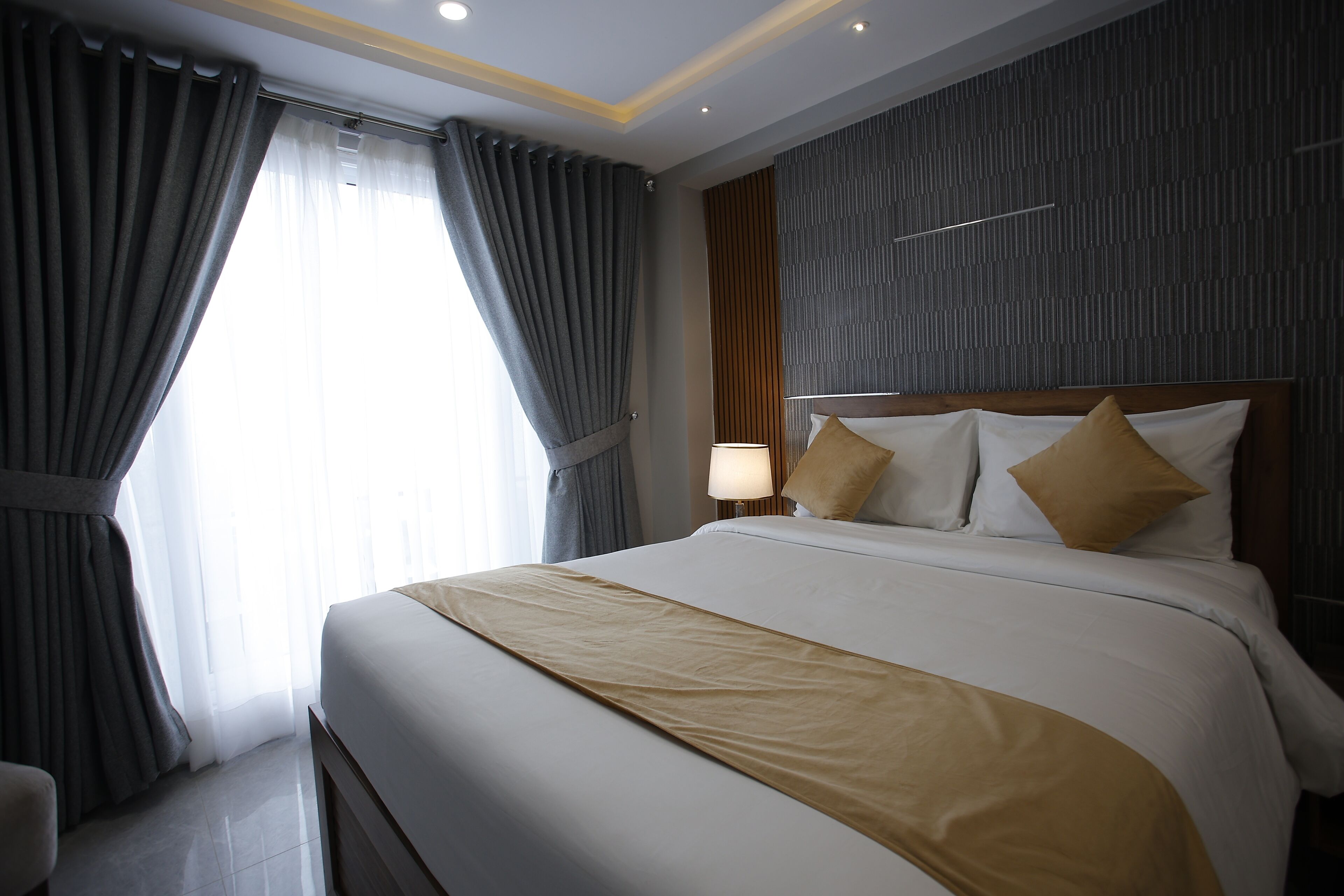 Habitación de hotel con una cama grande, una mesita de noche con lámpara y vistas al exterior a través de cortinas traslúcidas.