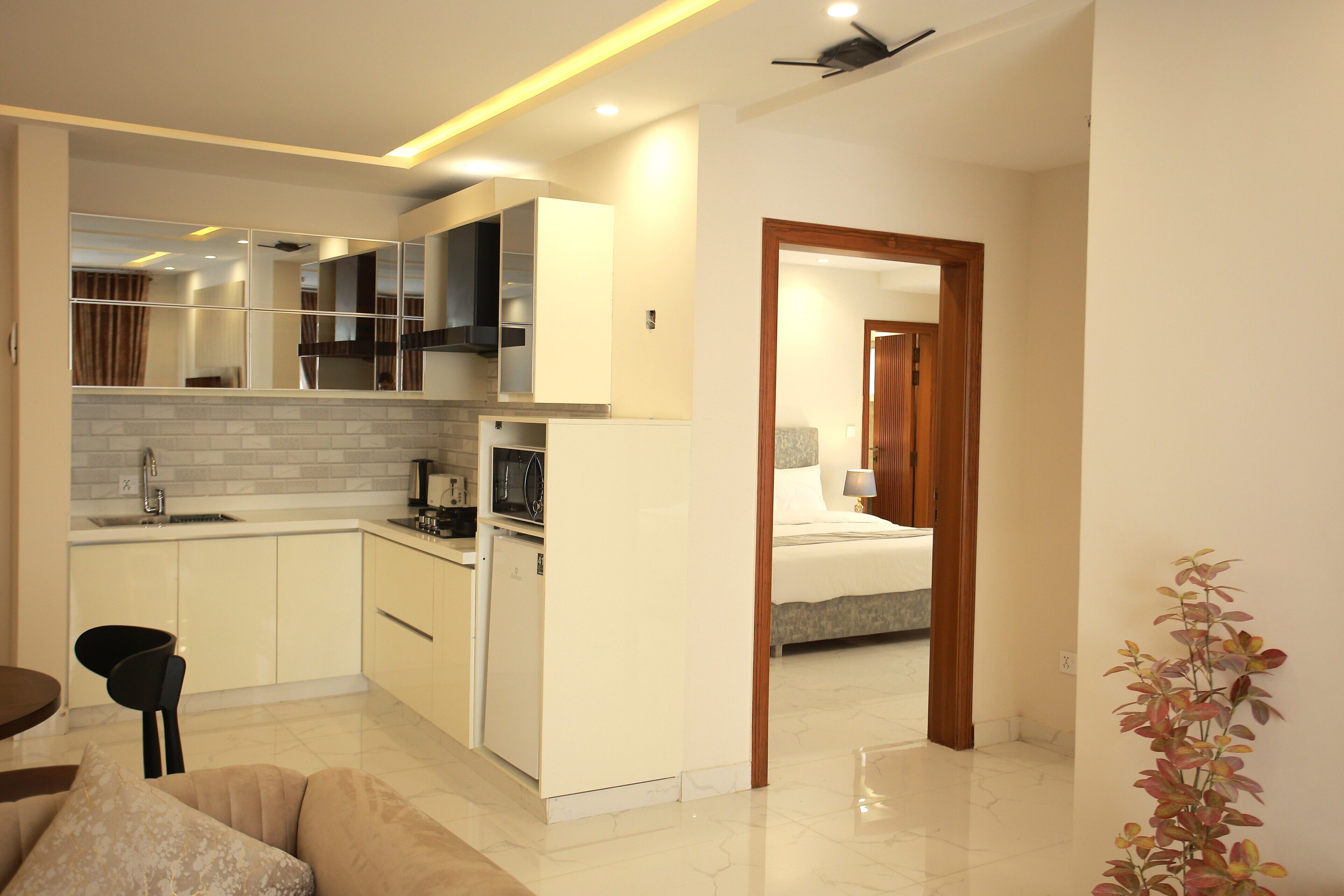 A modern kitchen with white cabinets, a built-in microwave, and a small refrigerator. There is a dining area with a black chair and a table. A bedroom with a bed and a nightstand is visible through an open doorway.