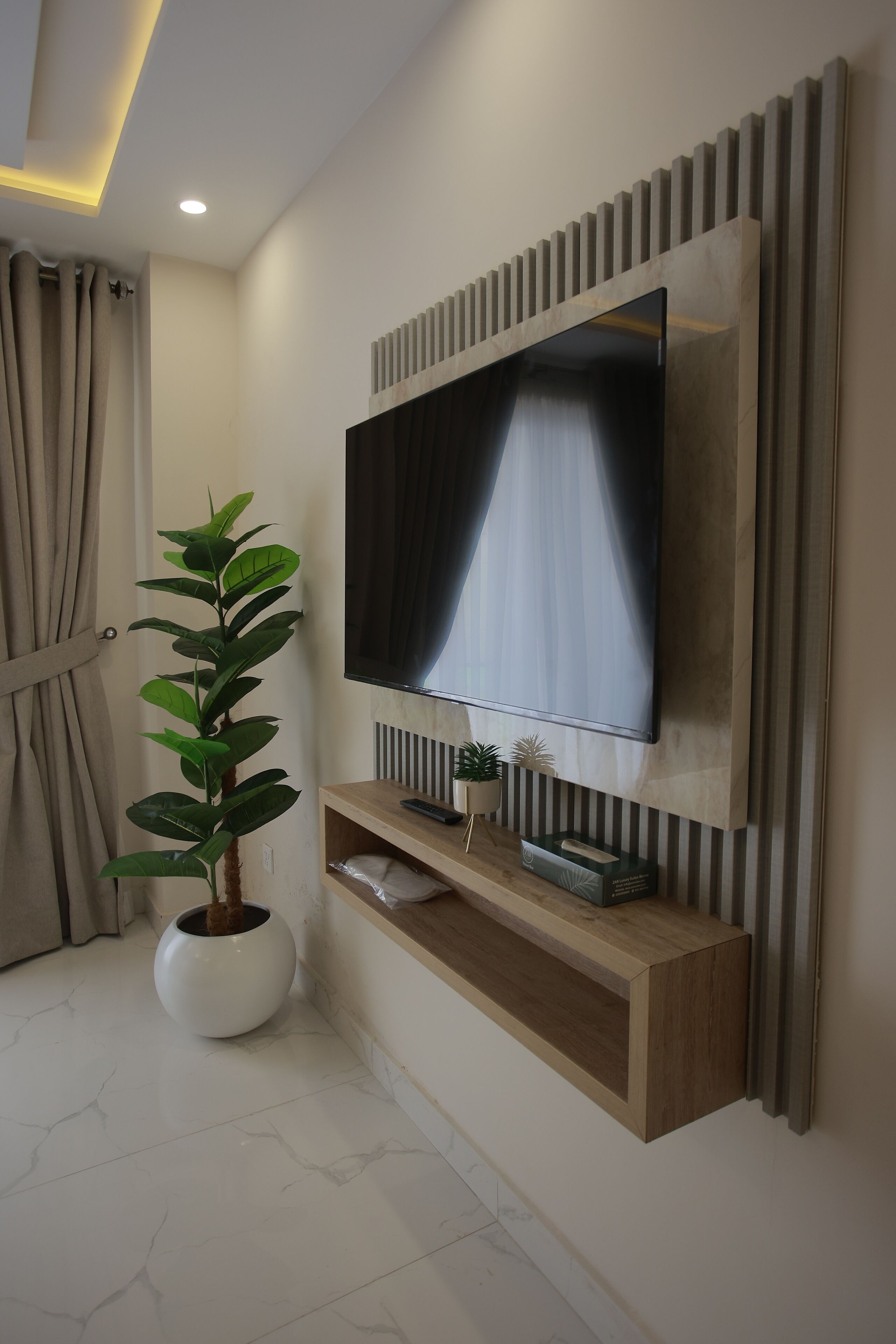 Ein modernes Wohnzimmer mit einem Wand-Fernseher, einem Holzregal darunter, einer Zimmerpflanze und einem Vorhang links.