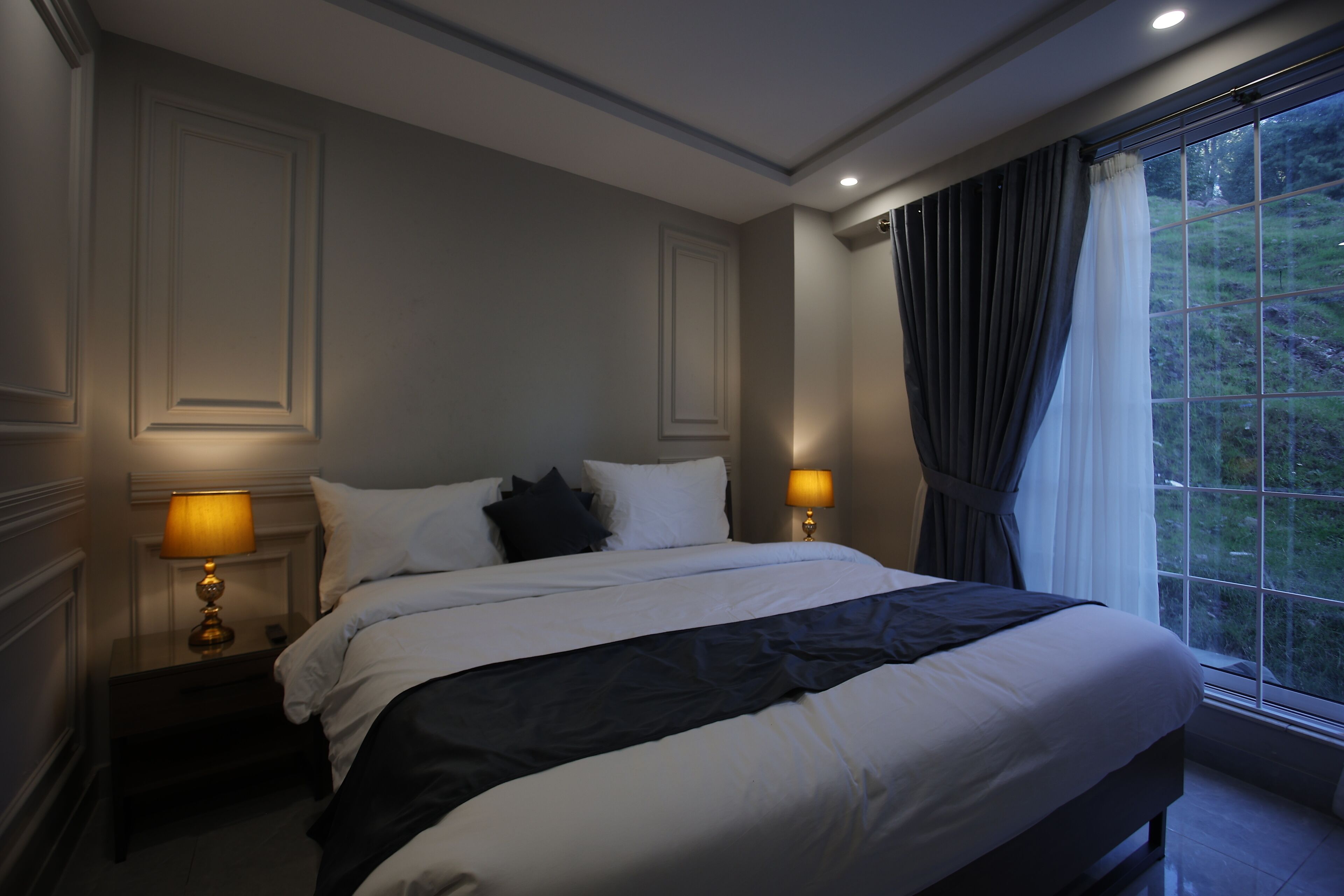 Uma cama bem arrumada com roupa de cama branca e preta, duas luminárias de cabeceira e uma janela com cortinas.