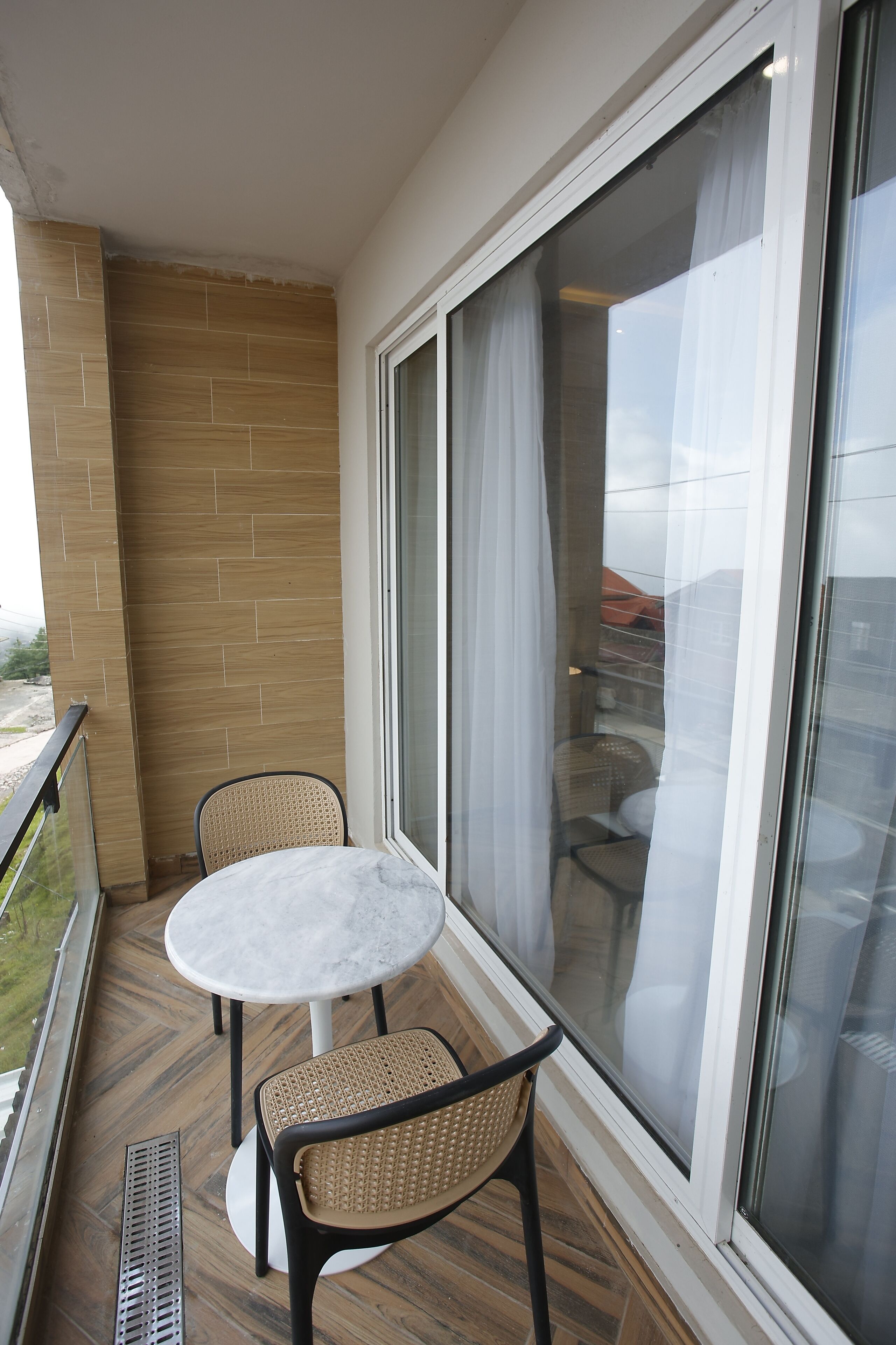 A balcony with a small table and two chairs, a large window with curtains, and a brick wall.