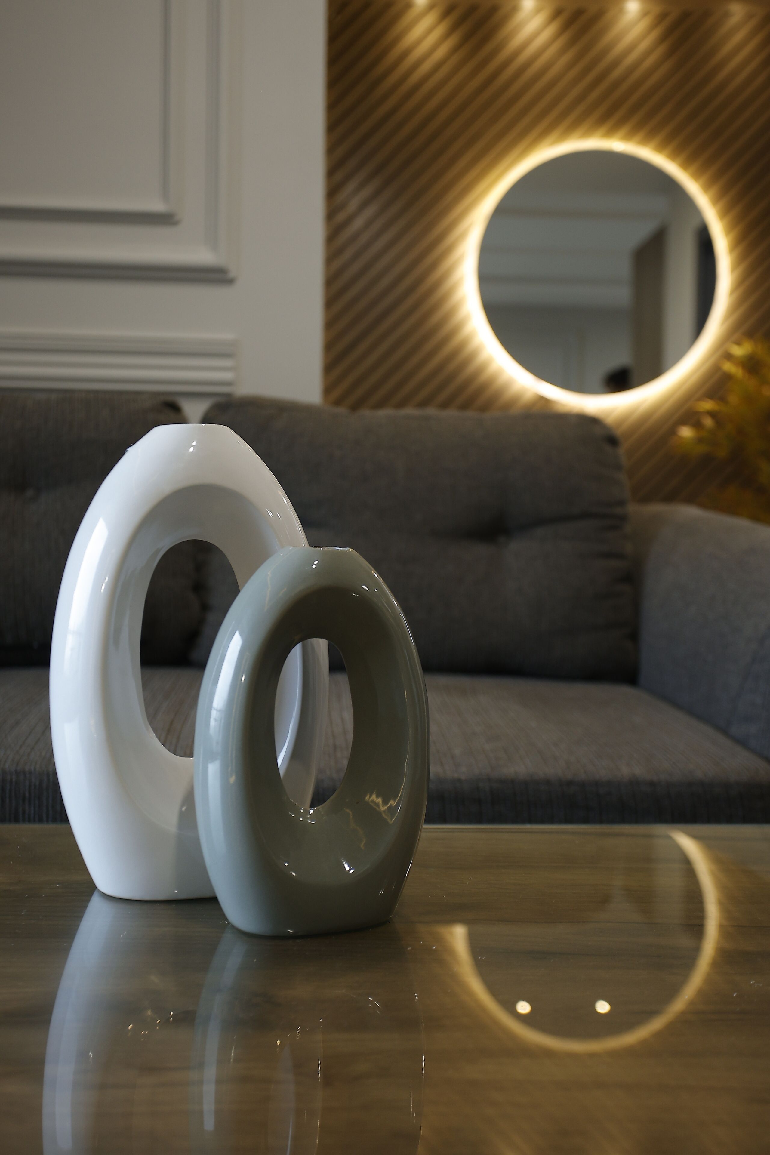 Two white and gray rings on a reflective surface in front of a gray sofa with a wooden wall and a circular mirror in the background.