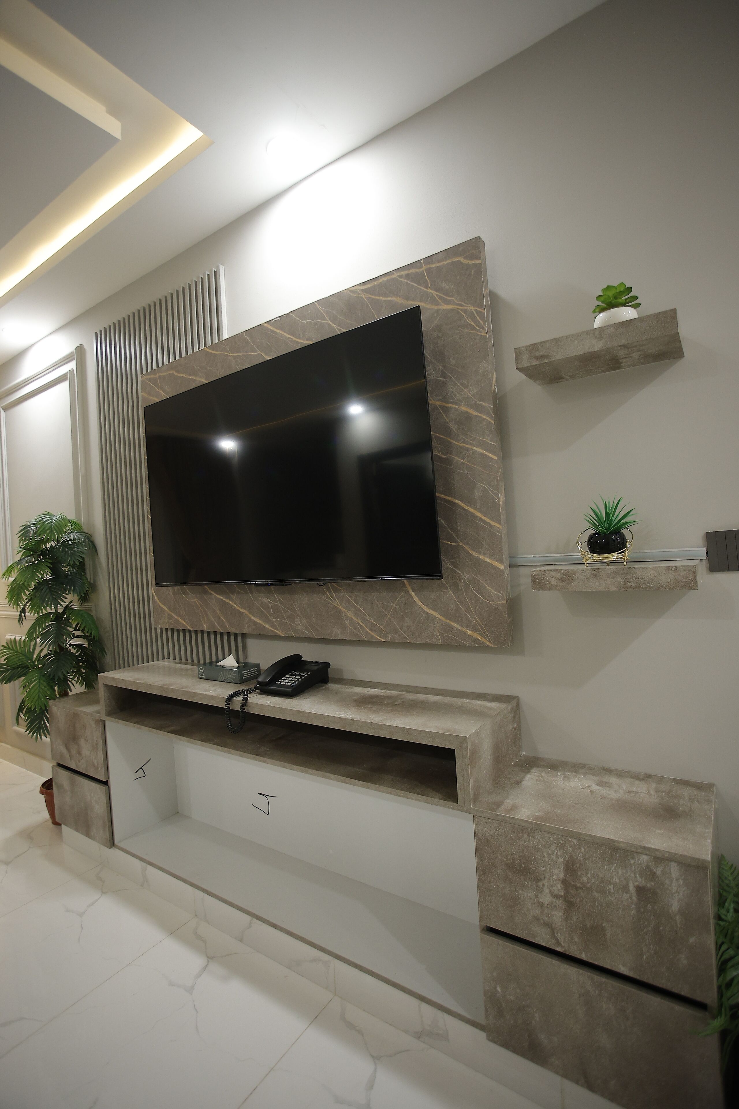 A modern living room with a large flat-screen TV mounted on a marble wall, a built-in sound system, and a small potted plant on a shelf.