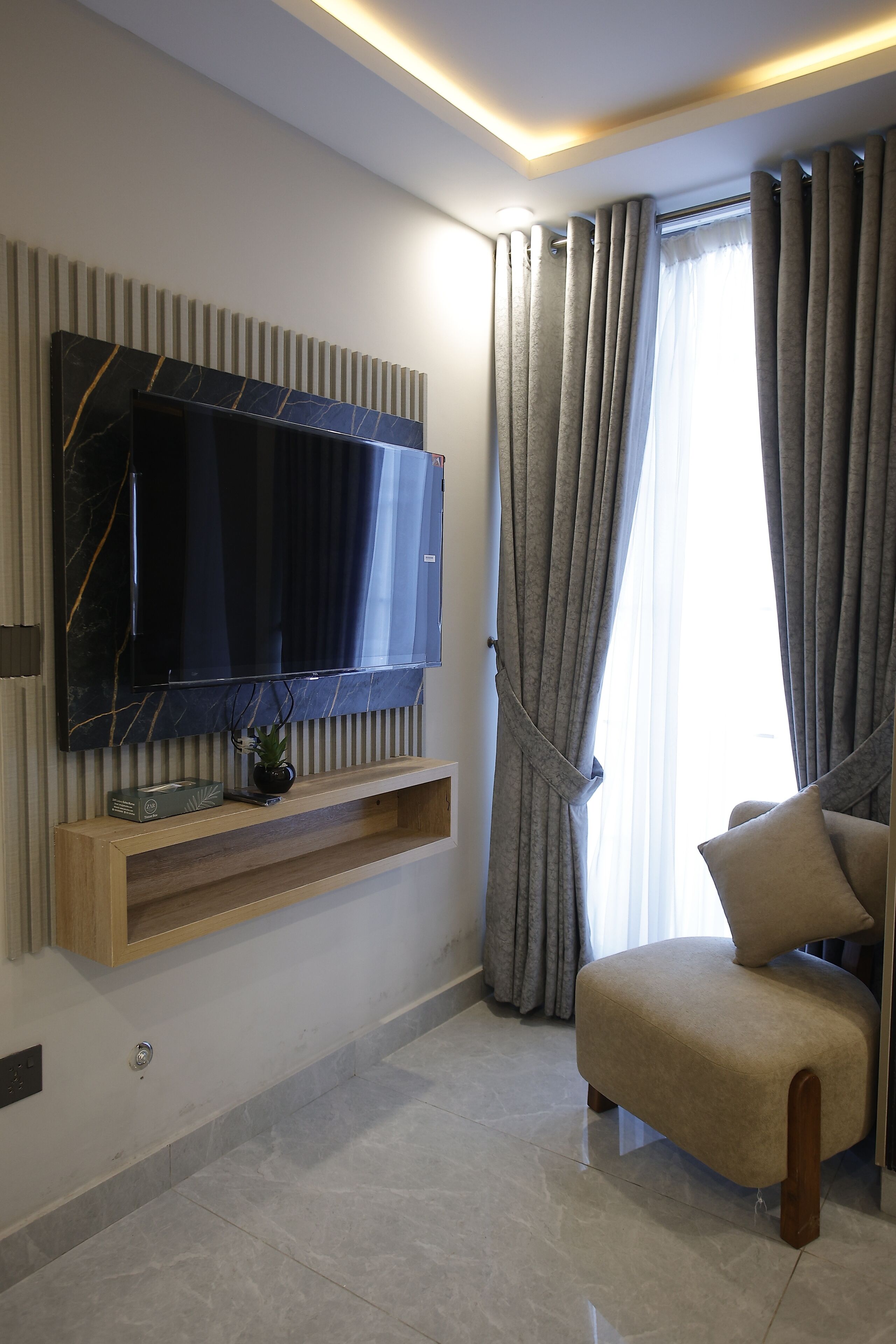 A modern living room with a flat-screen TV mounted on a wall, a wooden shelf below, and a comfortable beige armchair.
