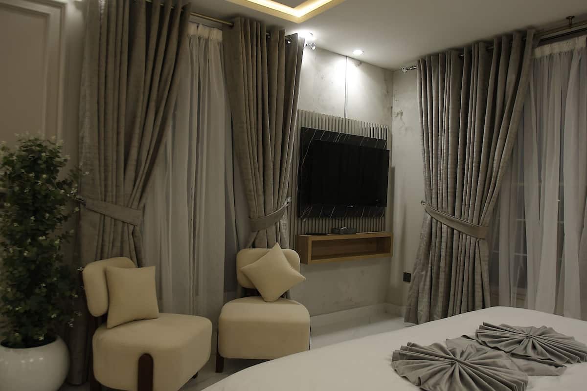 Une chambre à coucher moderne avec un grand lit, deux fauteuils, une plante en pot et un téléviseur fixé au mur.