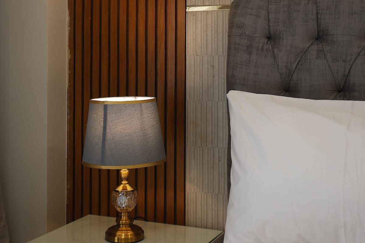 Un lit bien fait, avec du linge de lit blanc, une tête de lit capitonnée grise et une table de chevet avec une lampe.