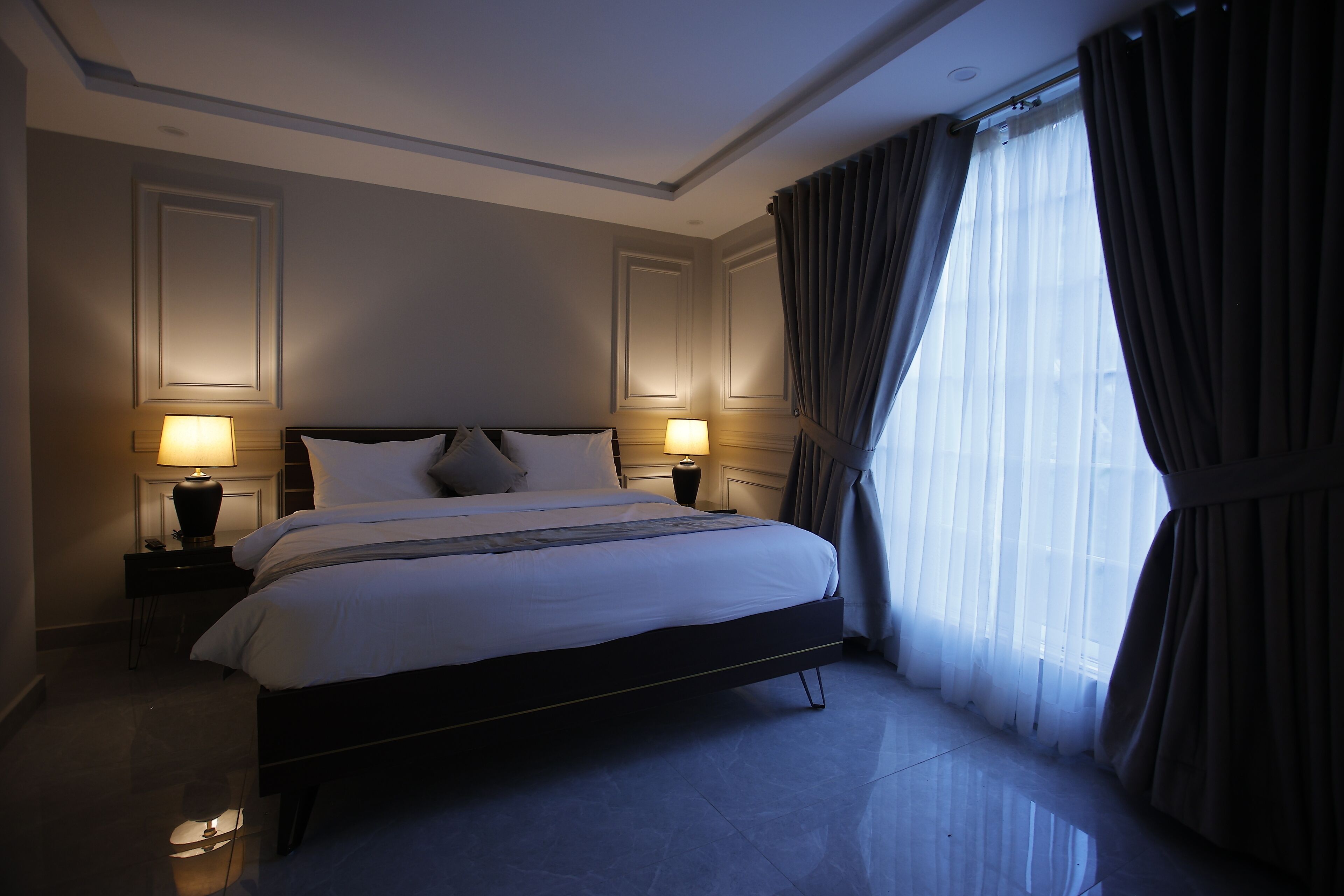 A hotel room with a large bed, two bedside lamps, a nightstand, and a window with curtains.