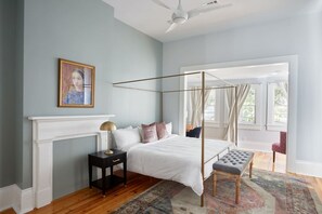 A bedroom with a four-poster bed, a nightstand, a chair, a fireplace, and a large window with curtains. - Spacious Magazine Apt | Walkable to Shops & Cafes (New Orleans)