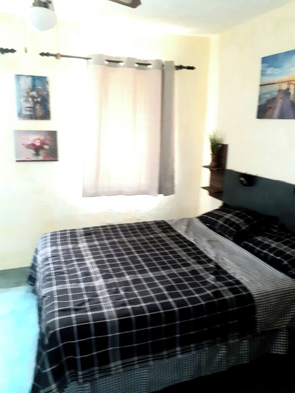 A bedroom with a bed, a window with curtains, and a wall-mounted shelf with a plant.