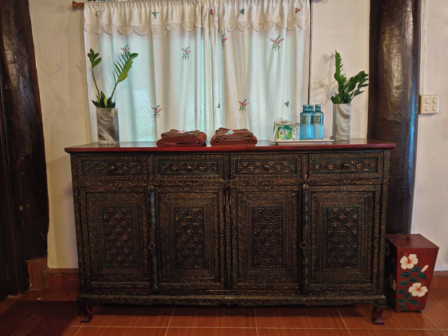 A dark wooden cabinet with intricate carvings, a window with floral curtains, and a small table with a vase of flowers.
