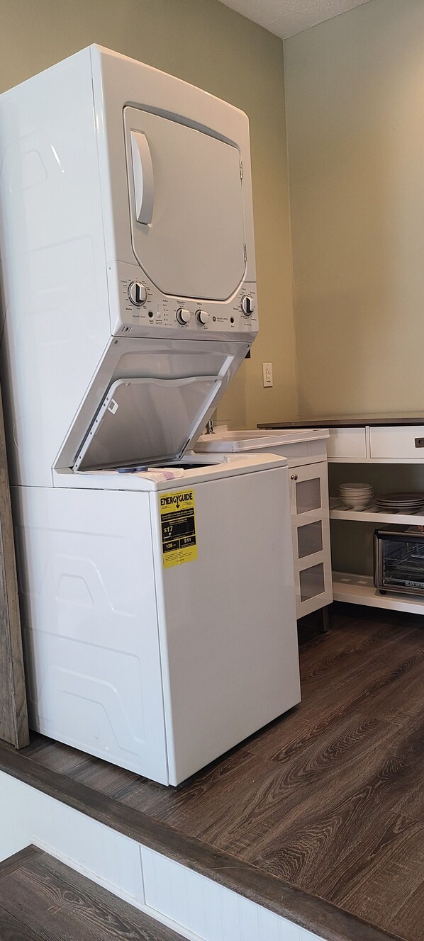 Una cocina compacta con lavadora y secadora apiladas, un microondas y una estantería con vajilla.