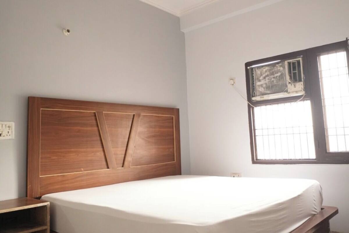 A bedroom with a wooden headboard, a bed with a white mattress, a ceiling fan, a window with bars, and a small wooden nightstand.